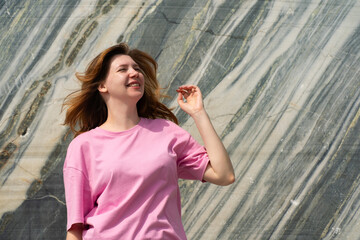 A cheerful young woman wearing a pink shirt poses with various expressions against a striking marble wall. Perfect for lifestyle, fashion, and casual themed projects. Captures joy, youth, and modern