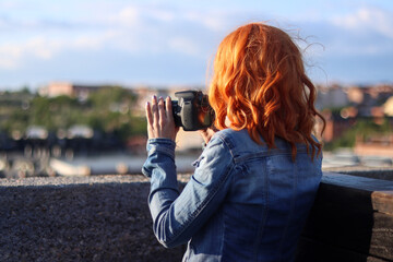 Young red haired woman using a photo camera at a viewpoint to photograph the city, image with copy space. Photography concept