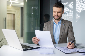 Smiling businessman working at desk with laptop, reviewing documents and taking notes in a modern office