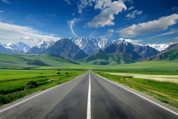 Fototapeta premium A wide road leads into the distance, with green fields on both sides and snow-capped mountains in the background. The blue sky is clear, and white clouds float across it