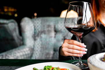 Close-up of a hand holding a glass of red wine against a blurred dining setting.