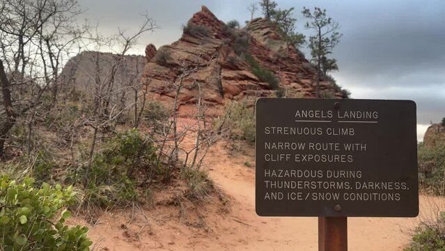 A warning sign marks the trailhead for the challenging Angels Landing Hike in Zion National Park, Utah. The iconic red rock formations and desert landscape of Zion Canyon loom in the background. 
