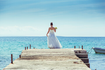 A bride stands at the end of a pier, gazing out at the tranquil, turquoise sea under a clear blue sky. The scene captures a serene moment of reflection and beauty by the water.