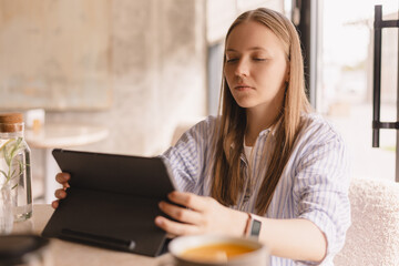 Fototapeta premium Attractive calm blonde woman wear shirt sitting with tablet computer in cafe. Beauty girl student using tablet computer for social media, studying or reading news.