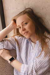 Portrait of blonde woman posing for portrait at home, sitting on comfortable armchair, looking with smile, laughing, leaning head on hand. Girl look sensual and dreaming wear shirt