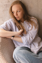 Fototapeta premium Portrait of blonde woman posing for portrait at home, sitting on comfortable armchair, looking with smile, laughing, fix her hair. Girl look sensual and dreaming wear shirt and jeans look side.