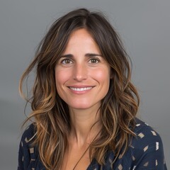 Fototapeta premium A woman with long, wavy brown hair and a warm smile wearing a dark patterned top, posing against a plain gray background in a portrait photograph