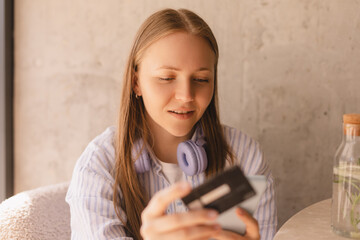 Calm young woman wear shirt with credit card in hand using mobile phone paying for purchase online relaxing sitting at table in cafe. Serious blonde girl make online order on the sale, shopping.