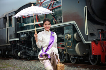 A portrait of a female in Thai traditional dress with umbrellas standing near a steam locomotive at the traditional train station.