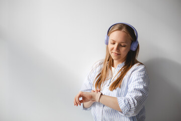 Checking time. Serious blonde worried girl wear shirt and listen music in headphones standing isolated white background looking at her watch while waiting for her friend. Happy woman read notification