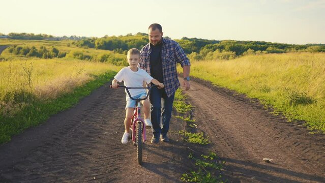 Happy little boy riding bike in summer vacation, father helping child balancing. Childhood and fatherhood moments, family weekend in nature, frontal view, active pastime in countryside in holidays