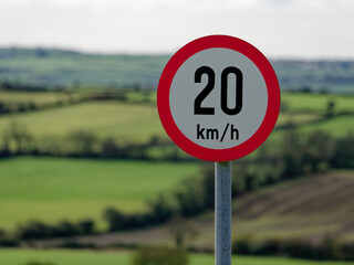A red and white 20 km/h speed limit sign stands against a blurred green backdrop, indicating a traffic regulation in a verdant area.