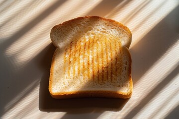A piece of toasted white bread in the shadow on the table. A slice of bread on a beige table