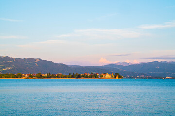 Germany, Warm orange sunset light shining on alps behind water of bodensee in summer with snow covered peaks next to old town of lindau city