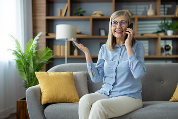 Fototapeta premium Cheerful elderly woman having a phone conversation while sitting on a comfortable couch in a cozy living room with modern decor.