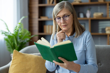 Senior woman with glasses sitting on a sofa, deeply engaged in reading a book at home.