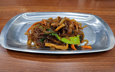 Delicious Korean Japchae Noodles with Vegetables on a Metal Tray, Photographed on a Wooden Table in a Casual Dining Setting