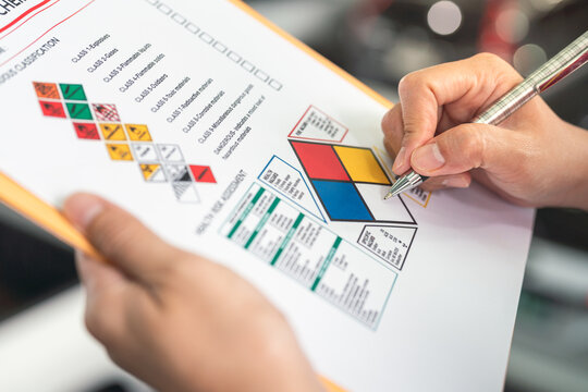 Action of a safety supervisor hand is checking on checklist of chemical and hazardous material form to verify the risk with blurred background of factory storage workplace. Industrial working scene.