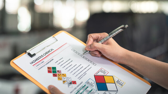 Action of a safety supervisor hand is checking on checklist of chemical and hazardous material form to verify the risk with blurred background of factory storage workplace. Industrial working scene.