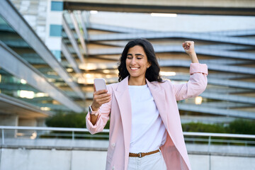 Exiting young middle eastern Israel businesswoman winner using smartphone mobile phone financial app. Happy smiling indian arabic woman in business suit celebrating yes win gesture holding cellphone