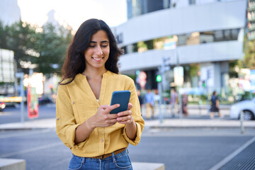 Smiling indian or arabic student woman in casual shirt holding cellphone for online business work or studying app. Young middle eastern Israel businesswoman using smartphone mobile phone outdoors