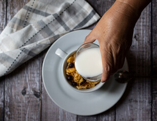 Concept of taking or grabbing food with hands, feminine Mexican hands. Serving milk over a white cereal cup on a rustic wooden table and a napkin on the side, horizontal flat lay or top view.