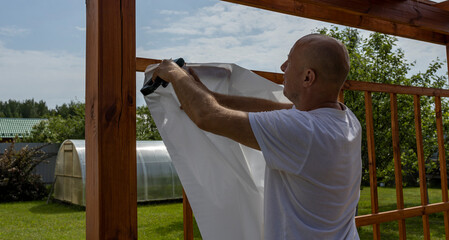 A man in a white shirt uses a tool to attach white fabric to a wooden pergola frame in a backyard setting.