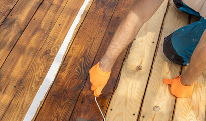 A person paints a wooden deck with a roller, leaving a line of unpainted wood. The sunlight shines...