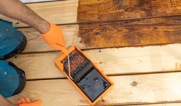 A person is staining a wooden deck with a dark brown stain. The deck is partially stained, with the top half of the image showing the stain applied and the bottom half showing the unstained wood.