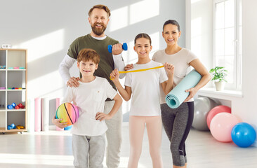 Portrait of happy smiling sporty family with kids standing in gym with sports equipment in hands...