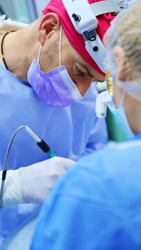 Focused male surgery professional wearing cap and headlight at operation. Doctor uses electric device. Close up. Vertical video.