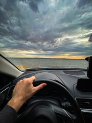 Scenic Drive by the Lakeside at Sunset: Hand on Steering Wheel, Overlooking Dramatic Clouds and...