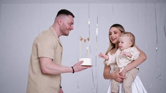 Caucasian mother wearing white dress holding a cute baby in hands. Father brings a cake and child stretches hands to it. Studio footage.