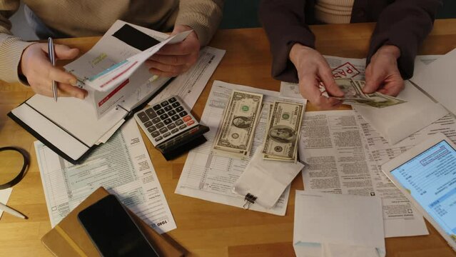 Top shot of hands of unrecognizable elderly female pensioner counting out American dollar bills and placing in three bundles on table, while husband is writing down utility charges in journal