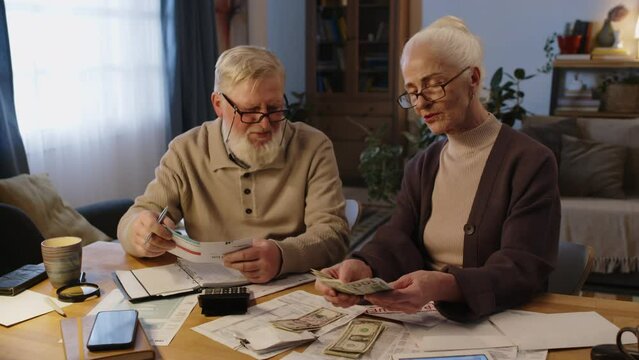 Medium arc shot of 70-year-old Caucasian lady in glasses counting US dollar bills, getting correct amount, talking to husband, who is studying utilities invoice, reacting with happiness and relief