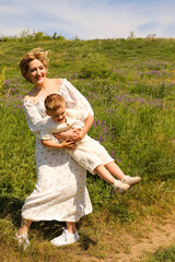 A woman in a white dress in summer holds a little boy under her arms and spins him around a green meadow.