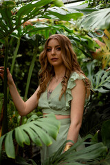  A woman with freckled hair and blue eyes is enveloped by large, green leaves, gazing intently into the camera