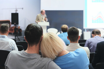 Fototapeta premium A man and a woman are hugging in a crowded room. The man is wearing a gray shirt and the woman is wearing a blue shirt. The room is filled with people, some of whom are sitting in chairs