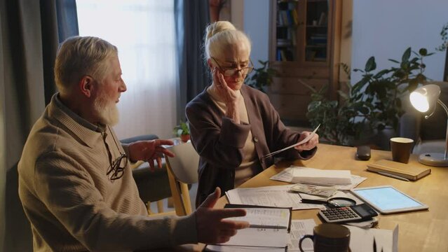 Medium shot of elderly Caucasian couple dealing with financial matters, woman taking overdue bill out of envelope, becoming upset and distressed, husband patting her on shoulder to calm down