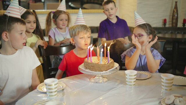 A birthday boy in a festive cap blowing out the candles on a birthday cake and his friends laughing and clap hands