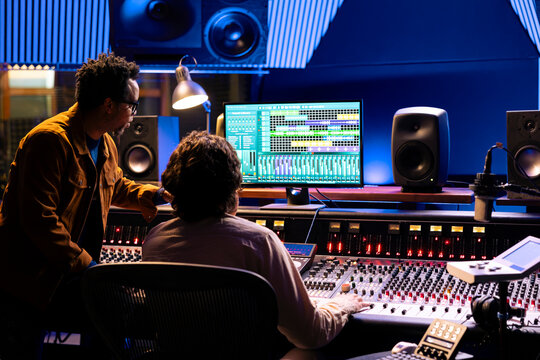 Singer and producer work on soundboard controller at recording studio station. Sound engineer uses digital console for music production, operating on faders and key buttons at control desk.