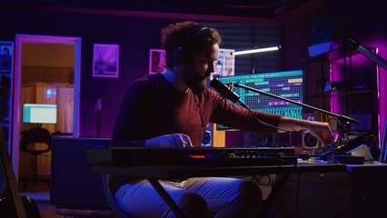 Sound engineer pressing keys of electric synthesizer and recording new music, adjusting noise level. Artist singing song on microphone, works with mixing board in home studio. Camera B.