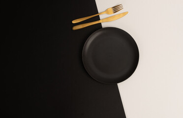 Fototapeta premium Black empty ceramic plate with gold cutlery on black white background. Center composition