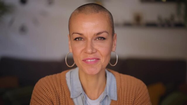 Cheerful attractive middle aged woman shaved hair looking smiling at camera. Joyful female people in their forties posing happy for portrait indoor with hoop earrings and positive aptitude at home