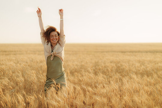 Happy woman with arms raised celebrating freedom and life in a beautiful golden wheat field at sunset, feeling positive and cheerful.