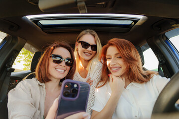 Three friends sharing a cheerful moment together while sitting in a car, capturing memories with a smartphone.