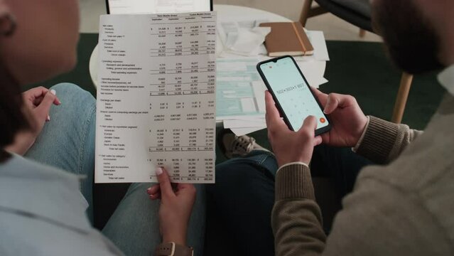 Over-shoulder shot of hands of young unrecognizable woman holding bank statement, pointing to numbers, and anonymous man adding them on calculator app on smartphone, while managing financial affairs