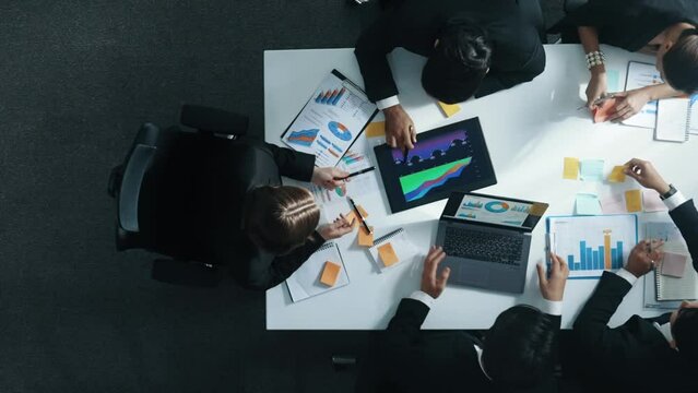 Top down aerial view of diverse business people discuss and brainstorm marketing idea while looking at financial graph at table with marketing document and sticky notes scatter around. Directorate.