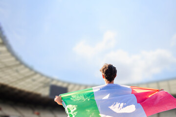 Passionate fan with the Italian flag draped over shoulders, standing proudly in front of a large...