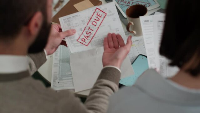 Medium over-shoulder shot of unrecognizable couple sitting at home, busy with financial matters, husband taking out overdue bill from envelope, ranting about high gas price, wife stroking his shoulder